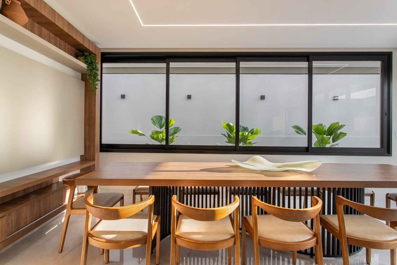Sala de jantar residencial com mesa de madeira maciça, esquadrias amplas e plantas — projeto de Cristiane Arquitetura